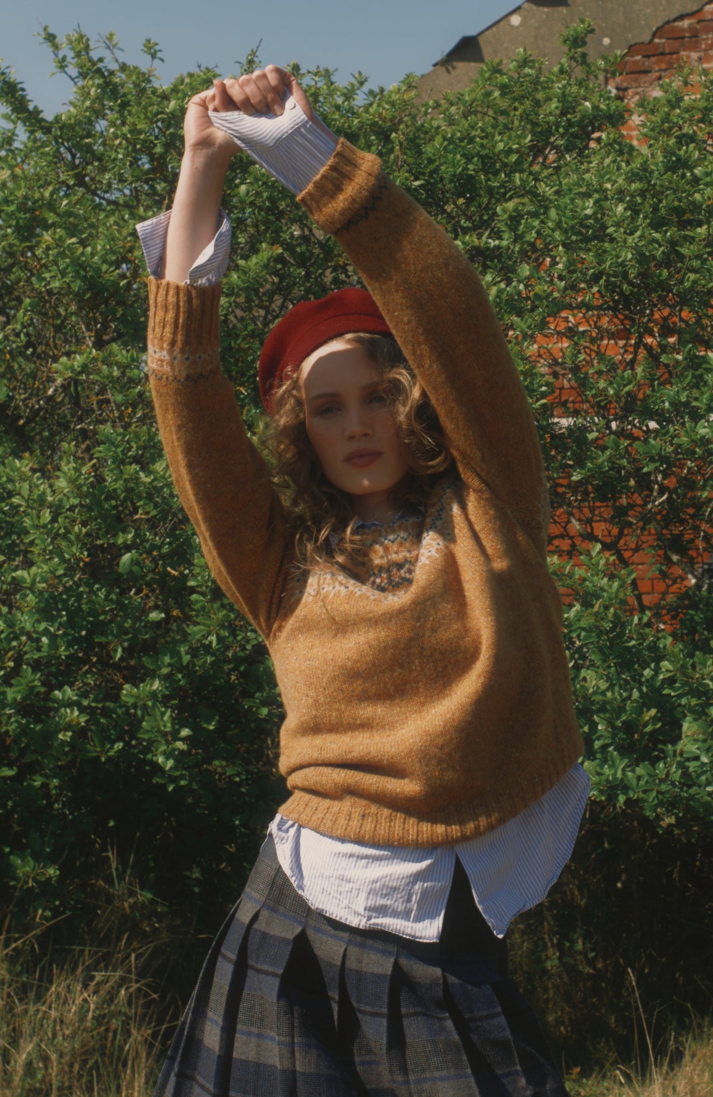 Person wearing a Bosie Islander sweater in Barra and plaid skirt standing in a field with greenery and a building in the background.