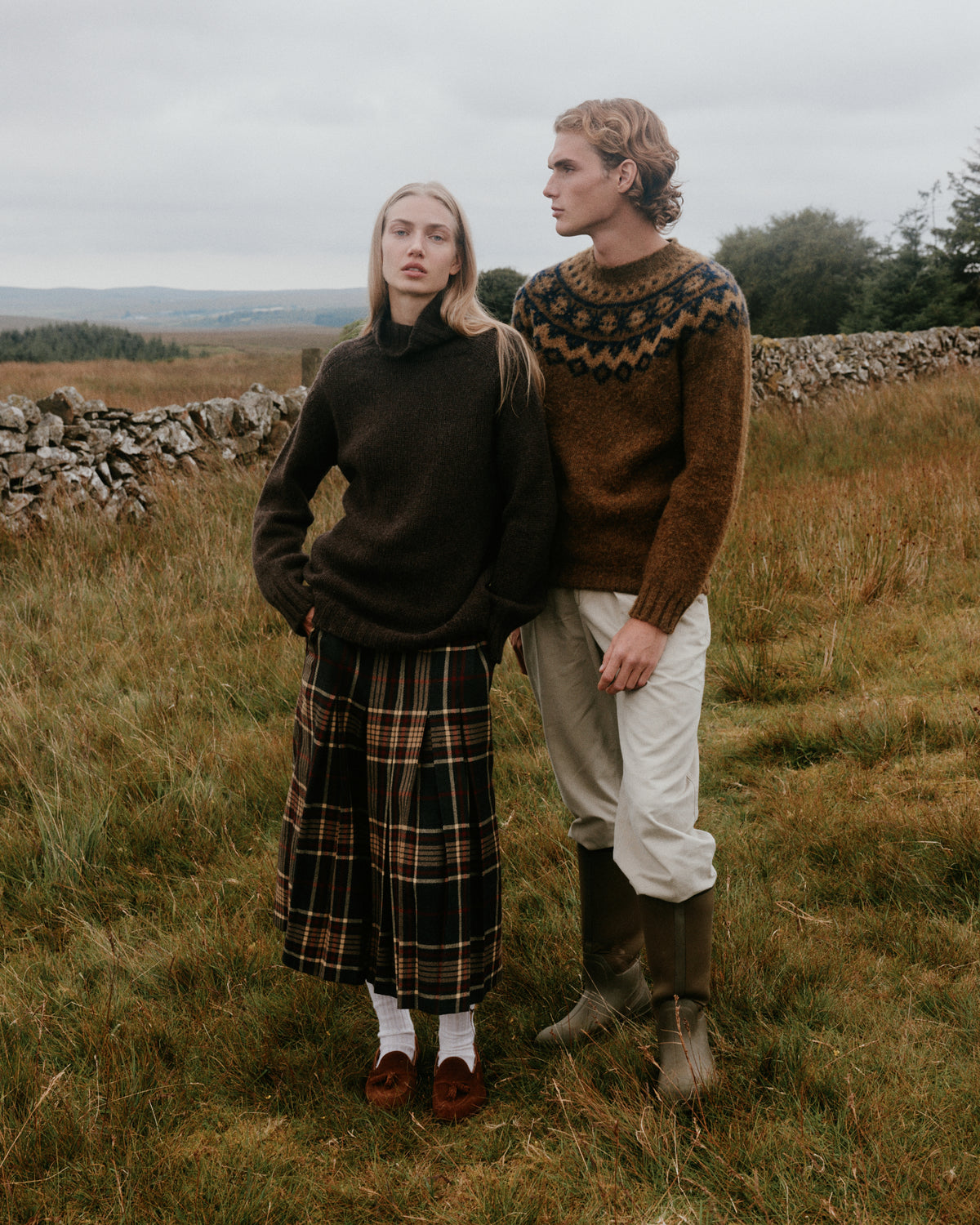 Woman in a dark brown roll neck jumper with a tartan skirt and man in a brown Fair Isle yoke sweater by a drystone wall on the Scottish moors – Bosie classic knits, made in Scotland