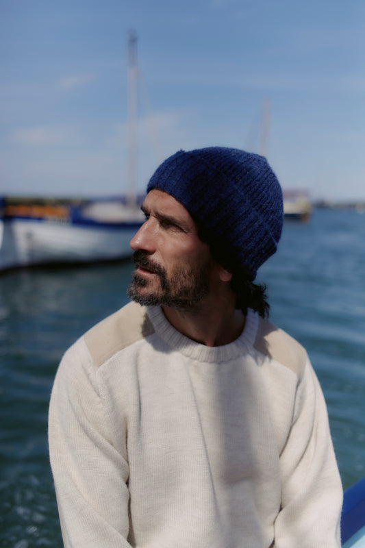 Man wearing a blue knit beanie and white sweater by a body of water with boats.