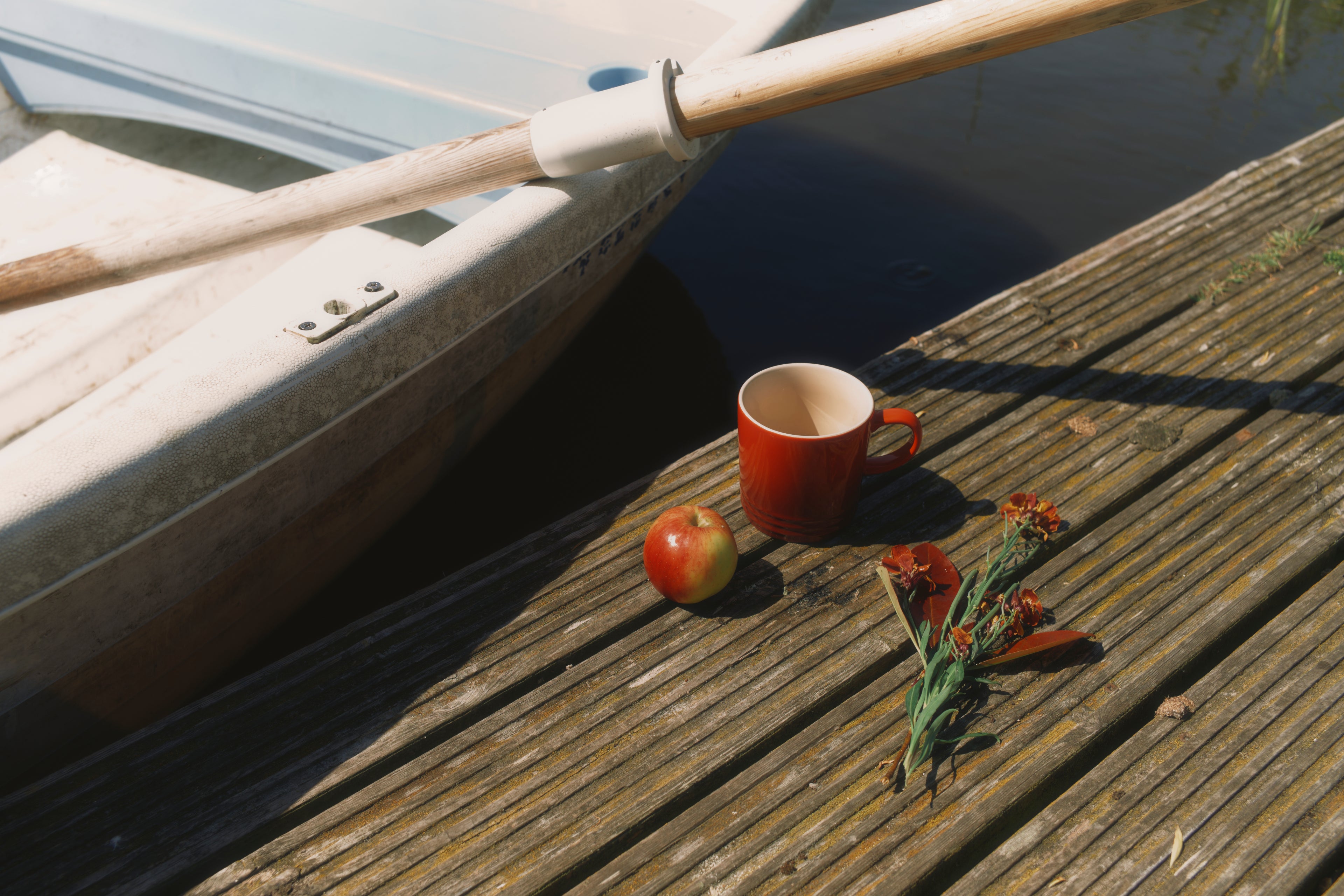Red mug, apple, and dried flowers on a wooden dock next to a boat.