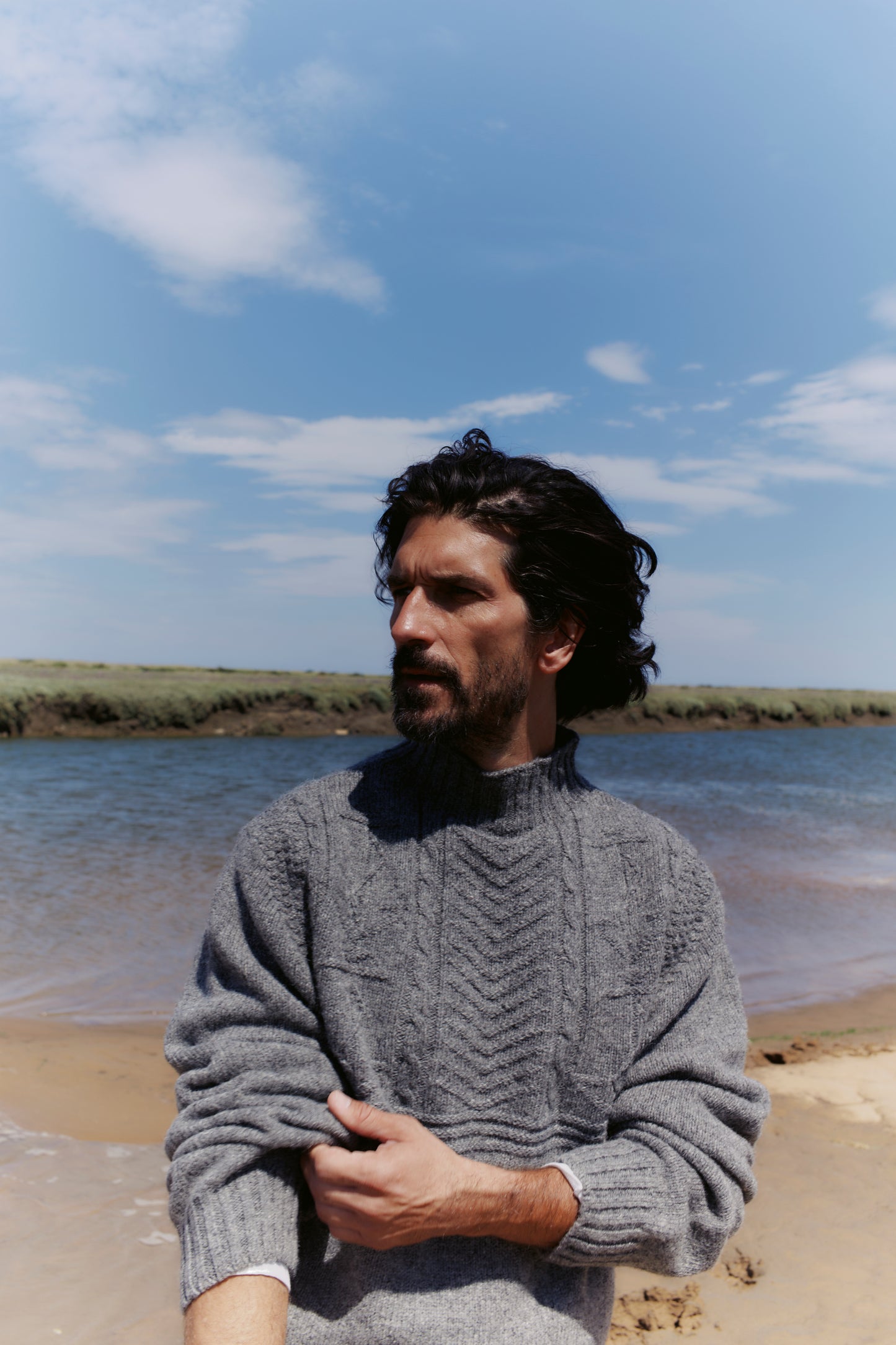 Man wearing a gray sweater standing on a beach with a blue sky and water in the background in a Bosie Ganjey.
