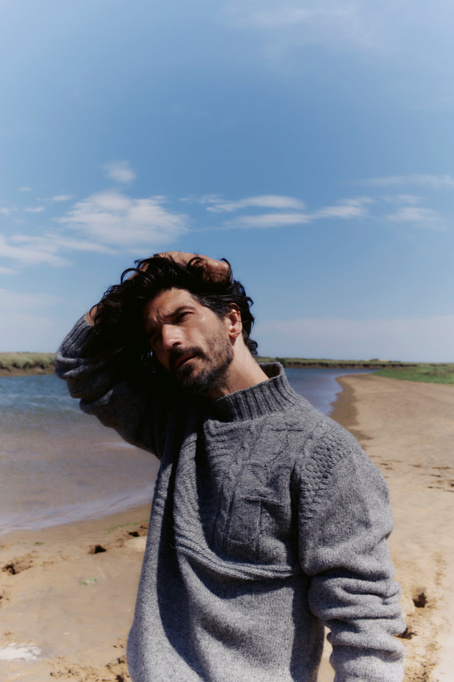 Man wearing a gray knitted Bosie Ganjey sweater by a body of water with a blue sky.