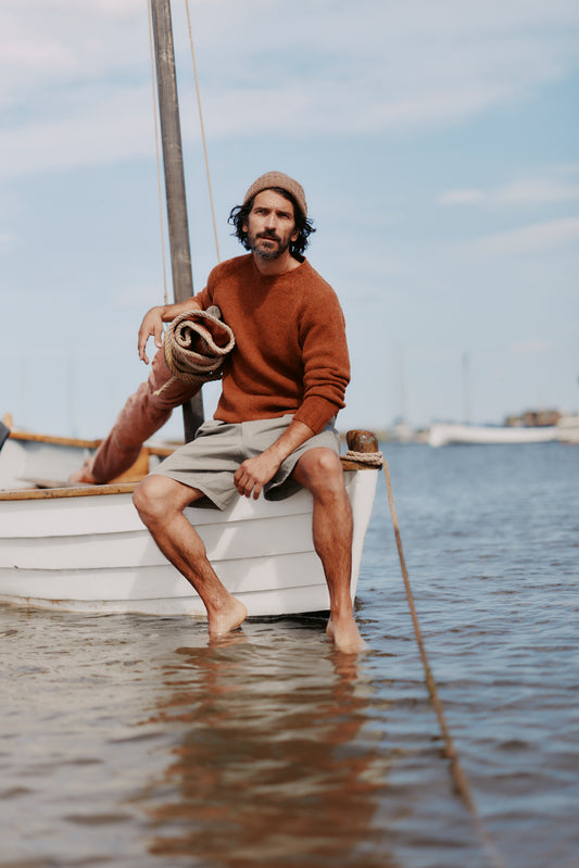 Man sitting on a small boat in water, Man sitting on a small boat in water, holding a rolled-up mat wearing a Bosie Knitwear Blue Mogganer North Sea Chunky Seamless Supersoft Shetland Sweater in colour Sienna Brown.