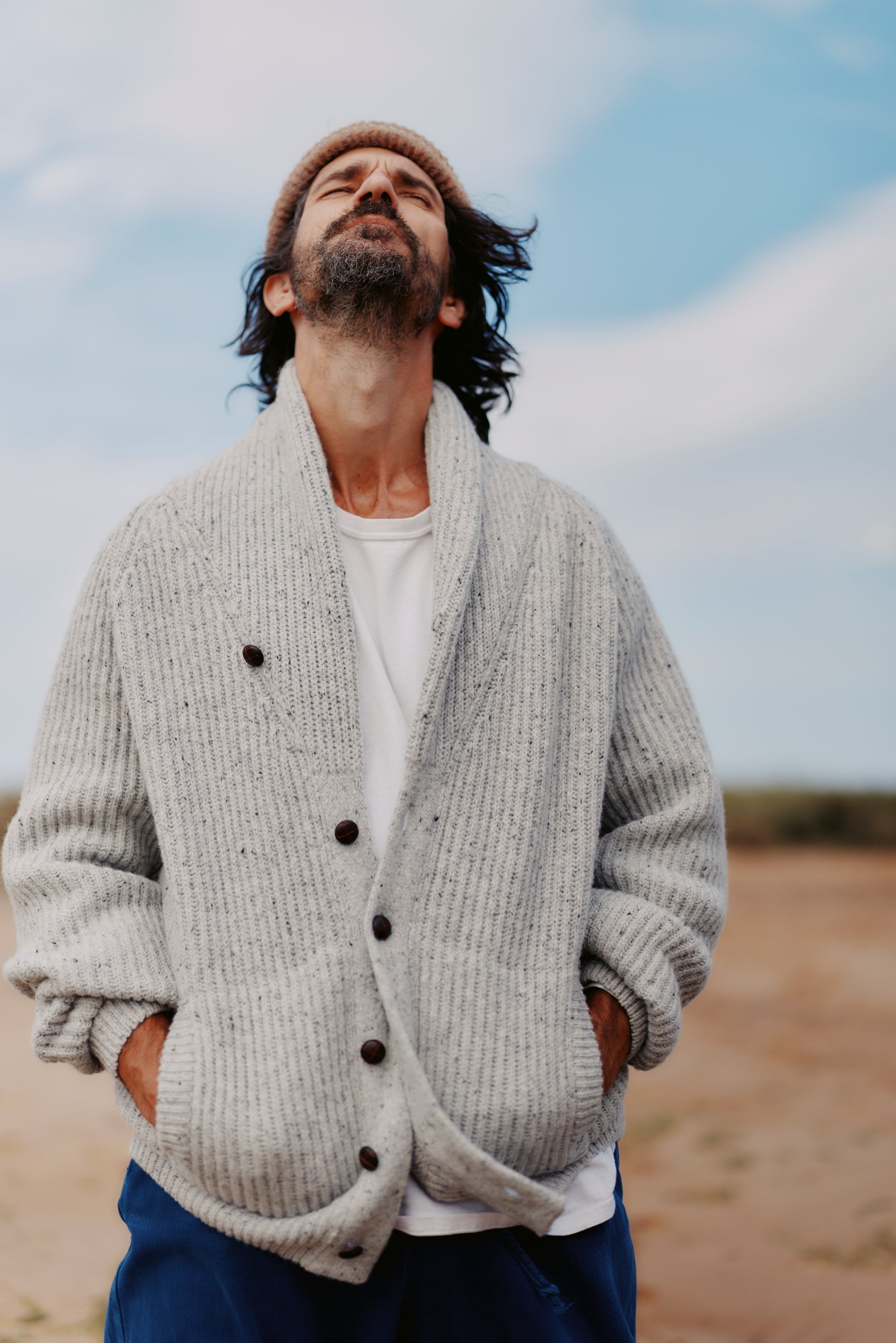 Man wearing a  Bosie Blue Mogganer Harbourmaster Cardigan over a white shirt with a beige beanie, standing outdoors with a blue sky and sandy ground.
