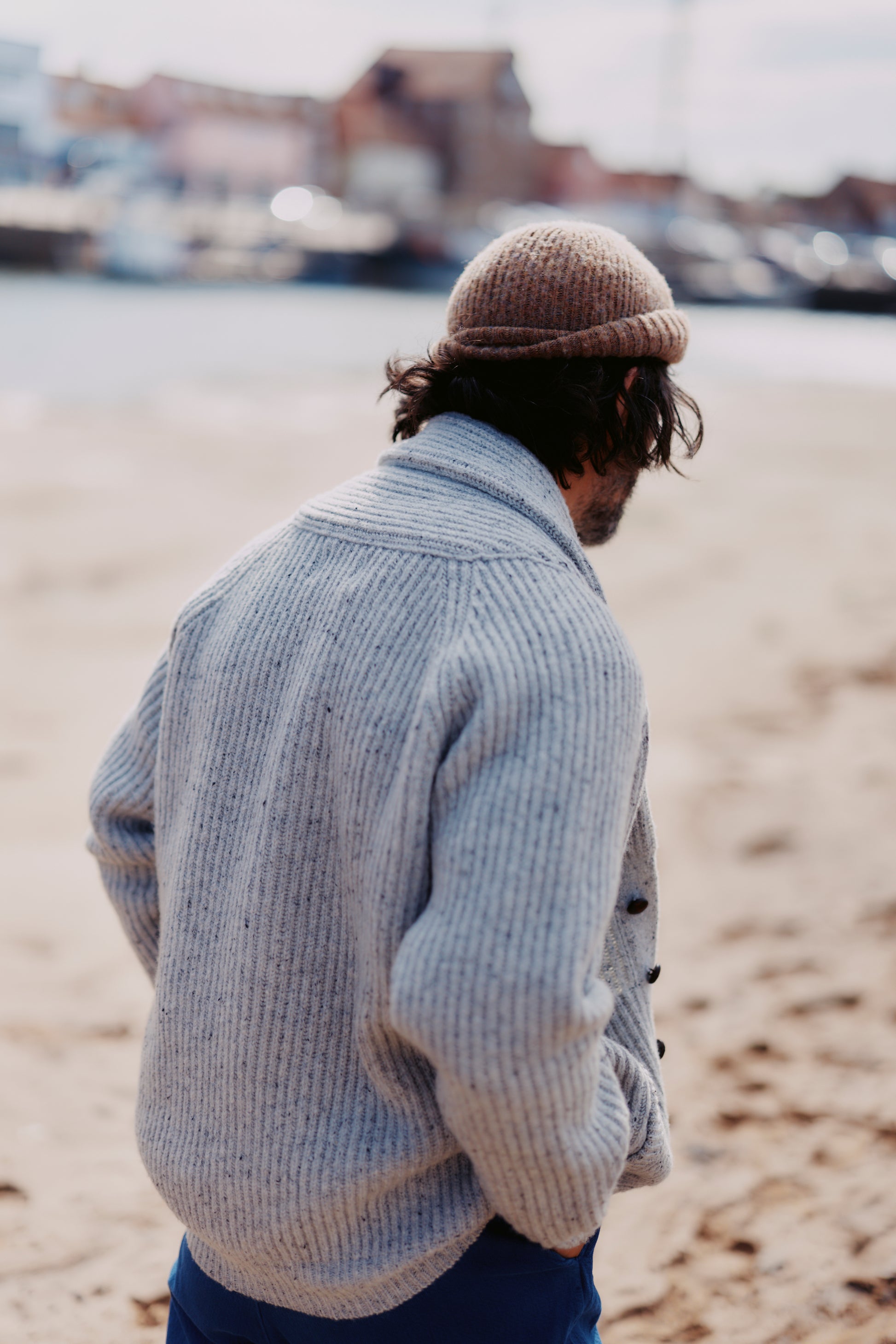 Man wearing a  Bosie Blue Mogganer Harbourmaster Cardigan and Heathland Brushed Hat  standing on a beach with blurred background