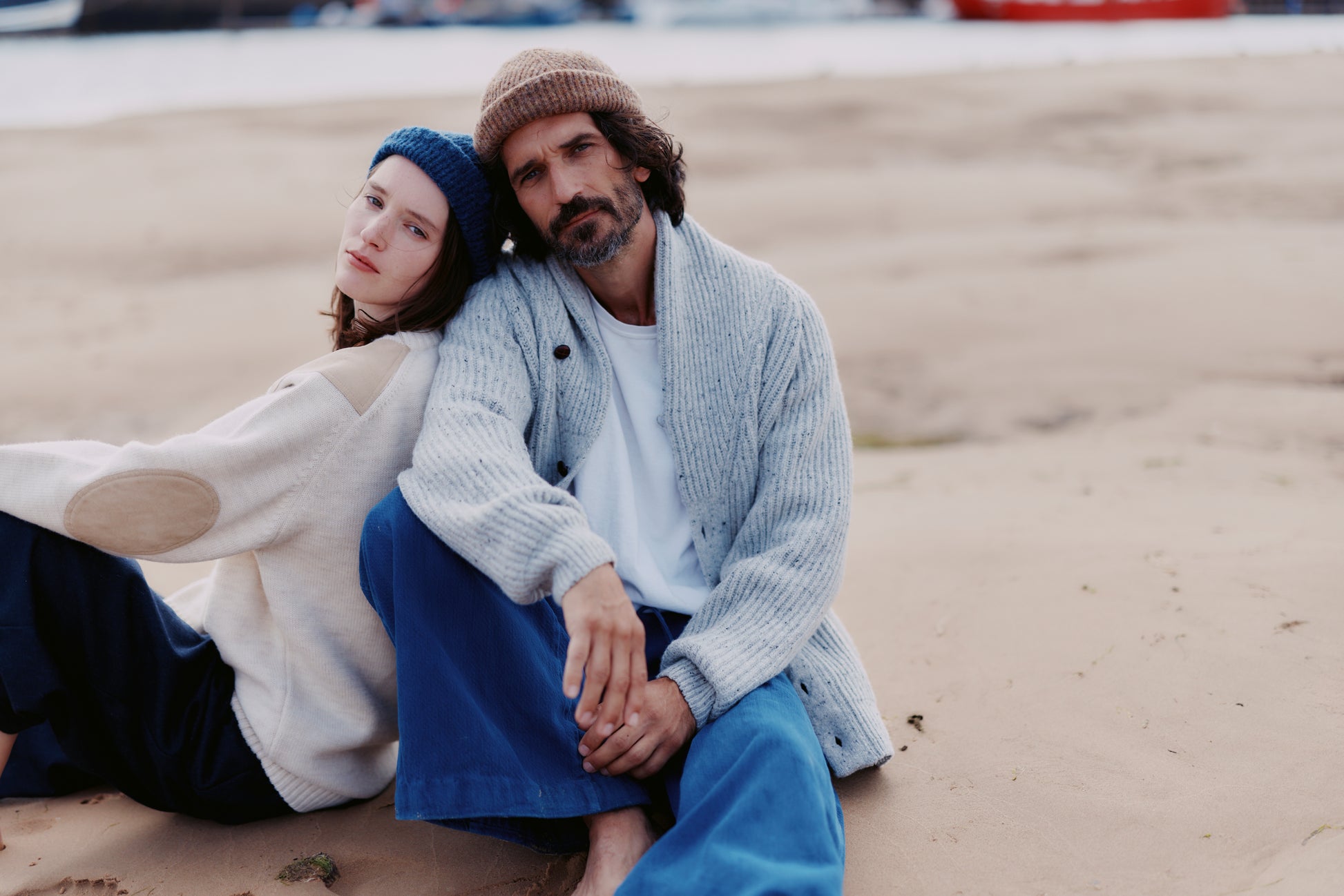 Two people sitting on a sandy beach wearing a  Bosie Blue Mogganer Harbourmaster Cardigan