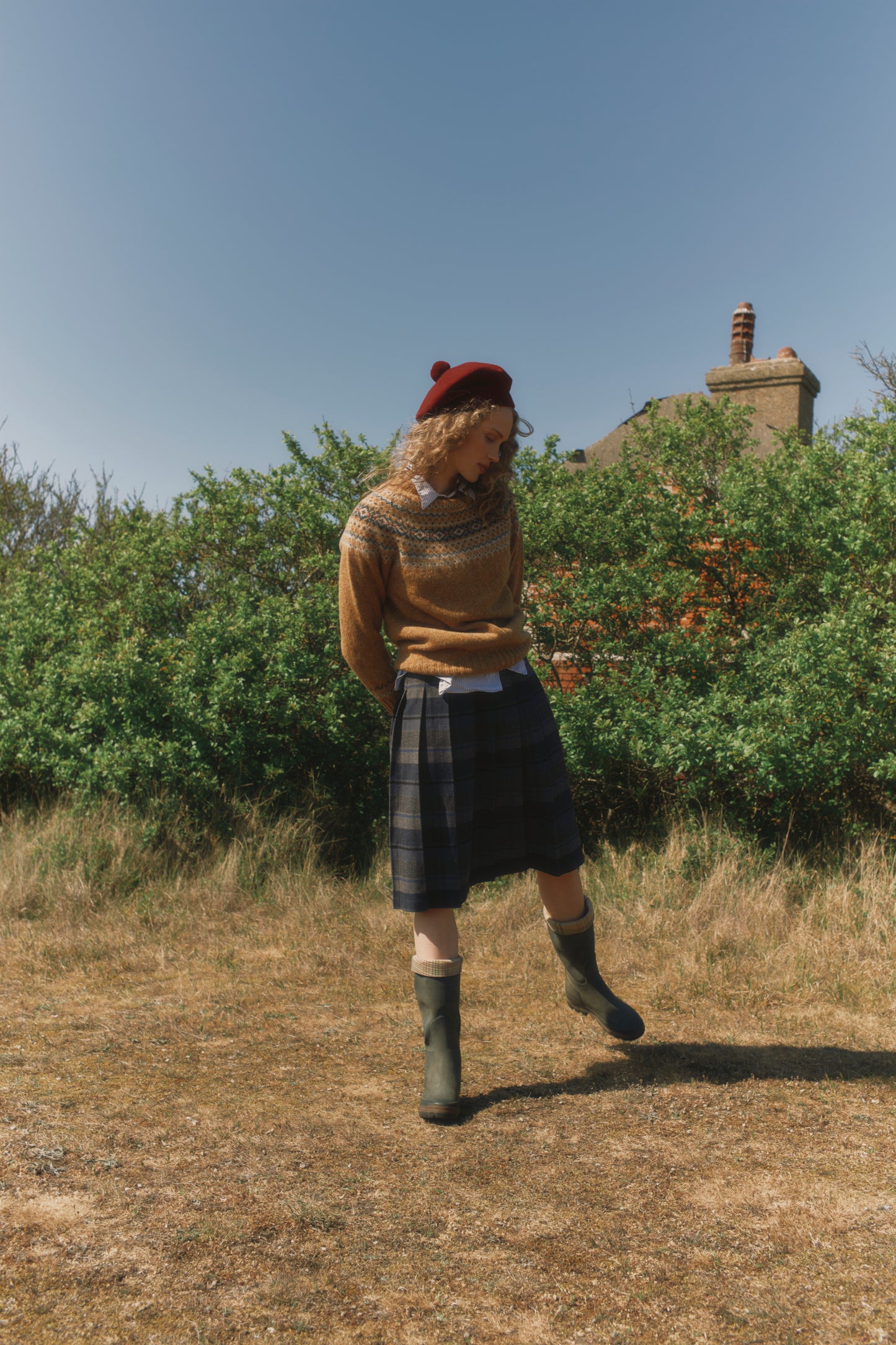 Person wearing a Bosie Islander sweater in Barra and plaid skirt standing in a field with greenery and a building in the background.