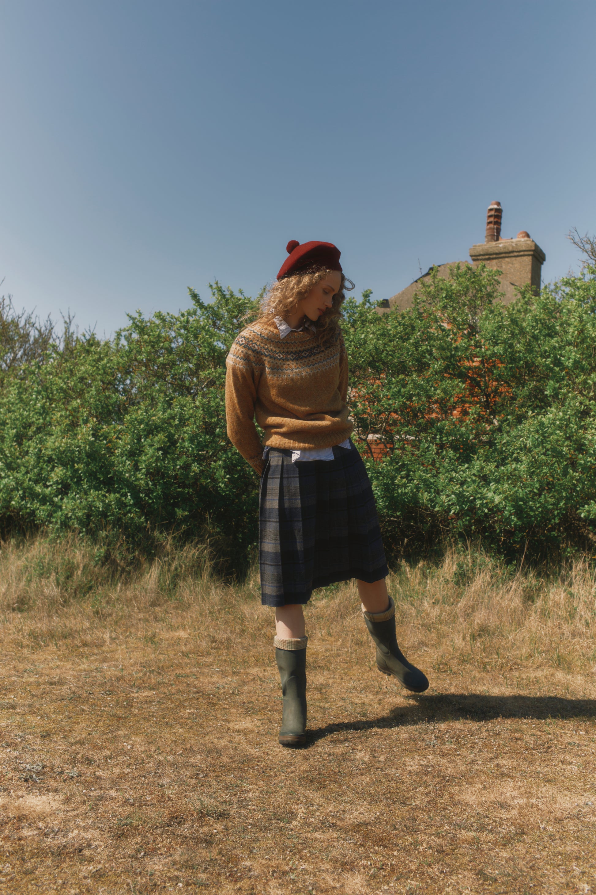 Person wearing a Bosie Islander sweater in Barra and plaid skirt standing in a field with greenery and a building in the background.