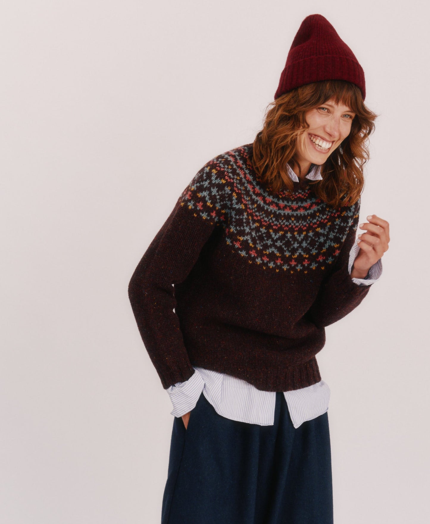 Person wearing a Bosie Luskentyre sweater in Lewis Dark Aubergine, navy pants, and a red beanie on a white background