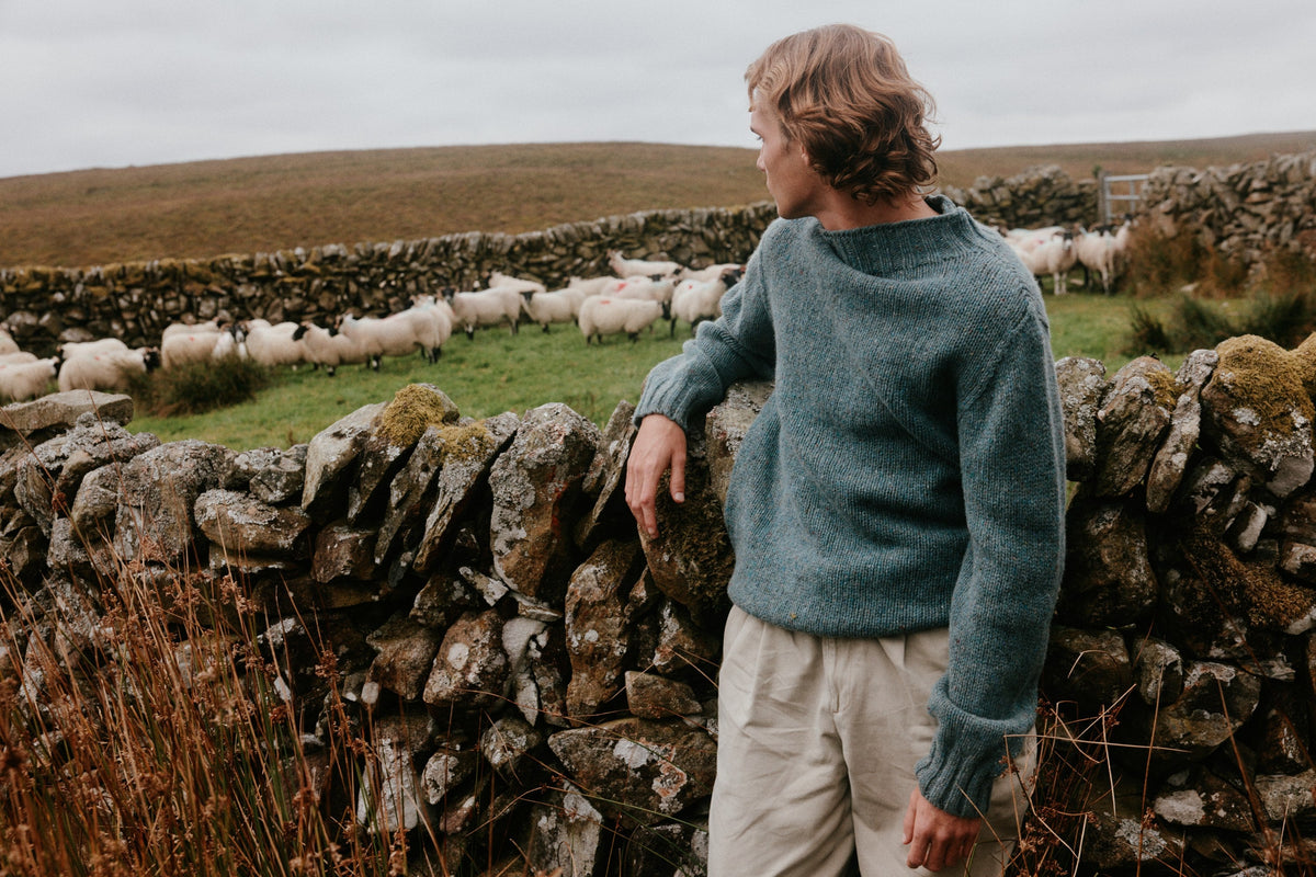 A man in a sea-green Bosie Lighthouse jumper leans on a drystone wall beside a flock of sheep in the Scottish hills – pure wool knitwear, knitted in Scotland