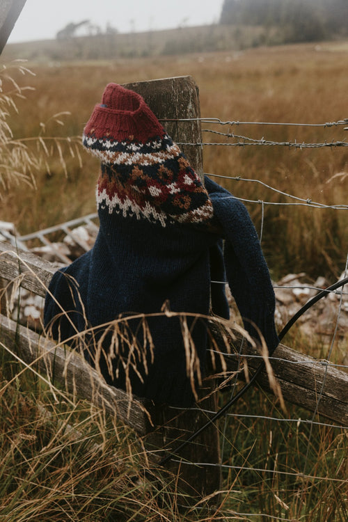 Navy wool jumper and red Fair Isle hat hung on a fence post in long autumn grass – Bosie Knitwear yarns, knitted in Scotland